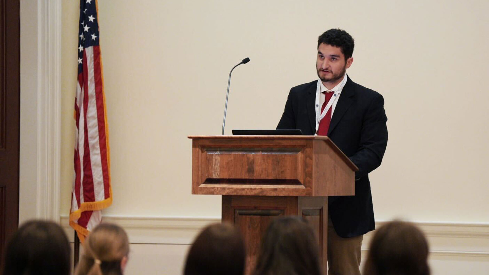 Man standing at a podium giving a lecture  with the American flag in the background 