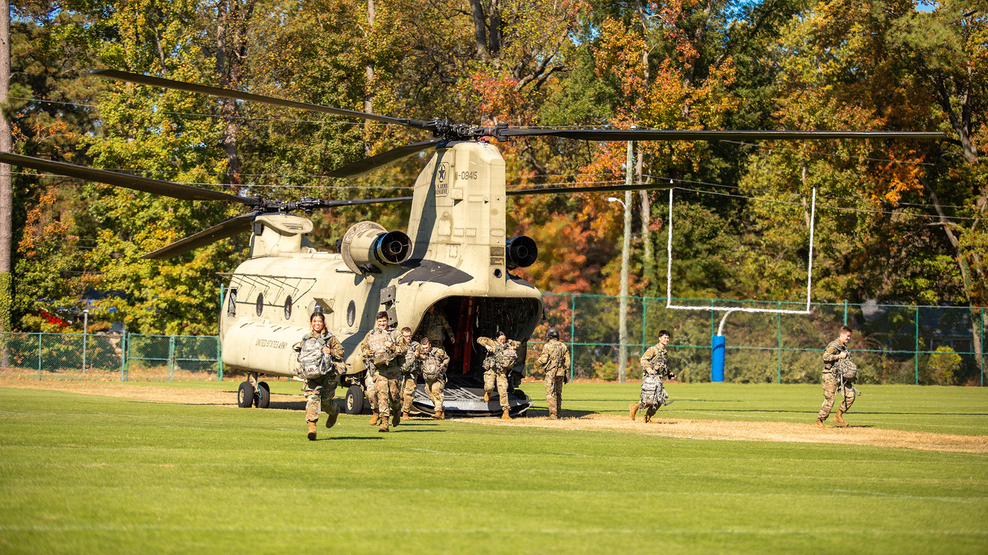 Army ROTC cadets run from a Chinook helicopter on a grassy field during a training exercise with autumn trees in the background.