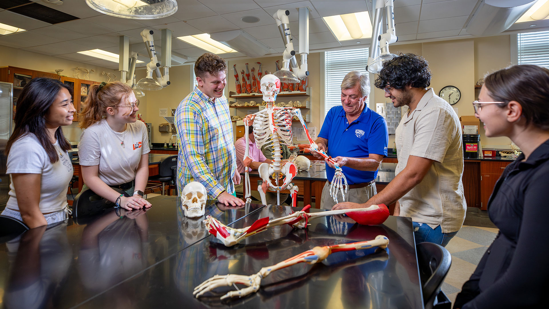 Students at a lab table with a model of a skeleton listen to a professor explain the musculoskeletal system