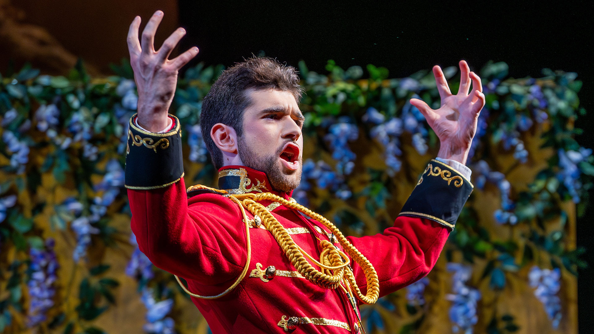 A man wearing a red military uniform holds his hands up in the air and flexed while singing