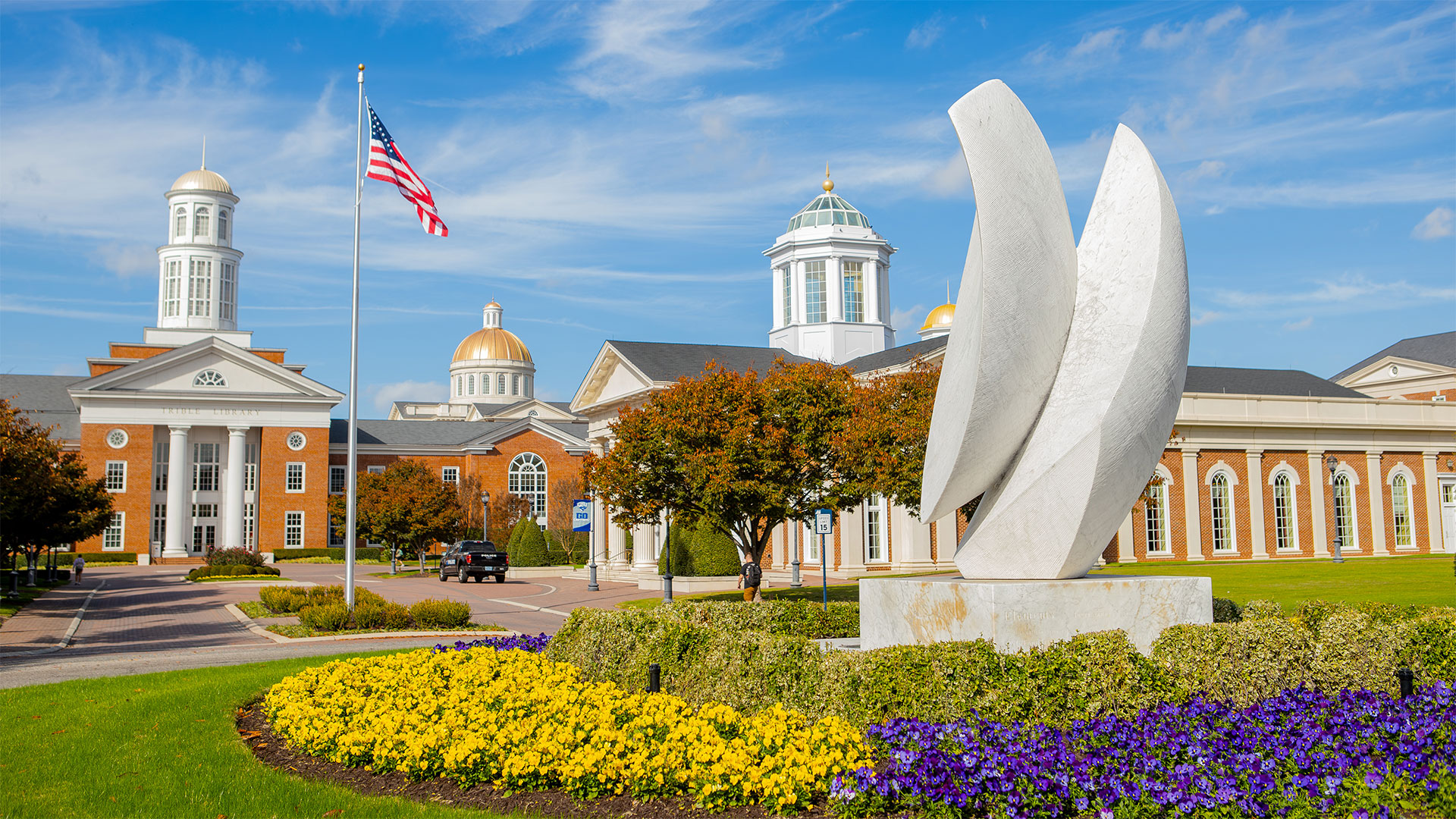The entrance of campus with a statue in the foreground and a flagpole with the American flag, the Trible Library and Pope Chapel in the background.