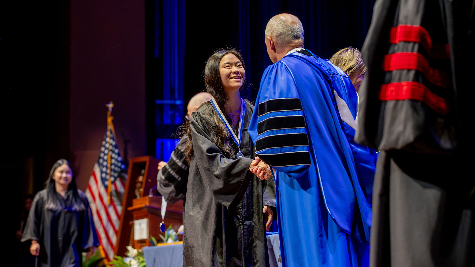 A student in a black graduation gown and a medal on a blue and white cord shakes the hand of University President Bill Kelly.