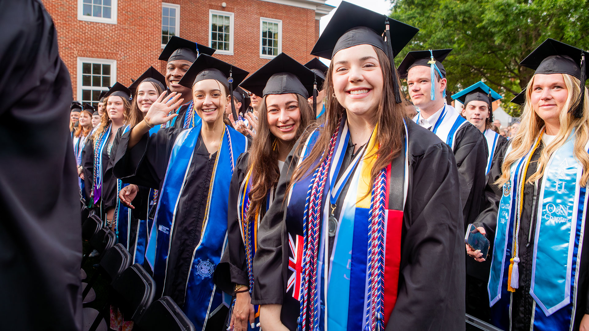 - A group of happy graduates in black regalia, multi-colored cords, and stoles smile and wave during commencement.