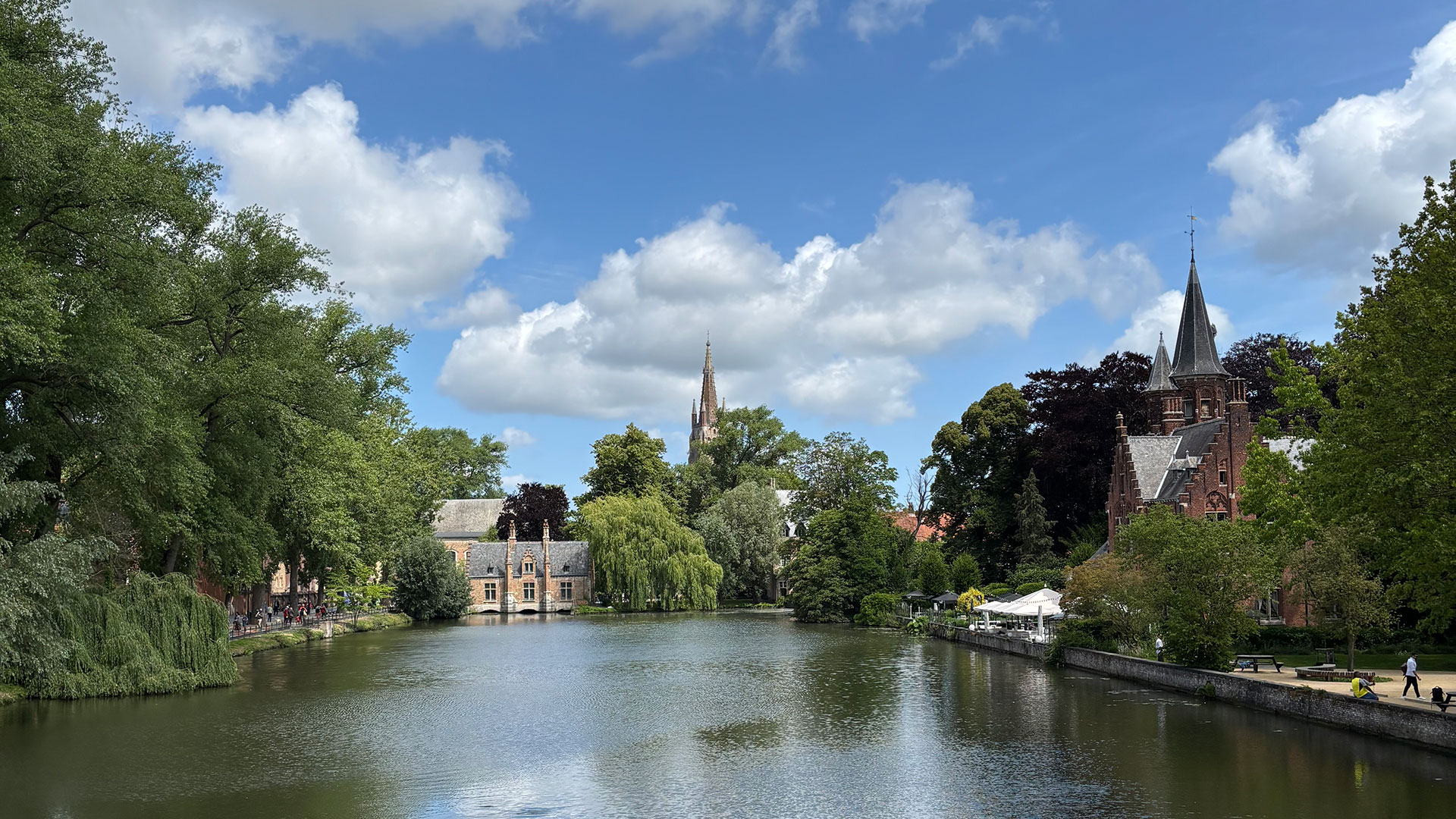 Scenic view of the Minnewater Lake in Bruges, Belgium, with historic brick buildings and lush green trees under a blue sky.