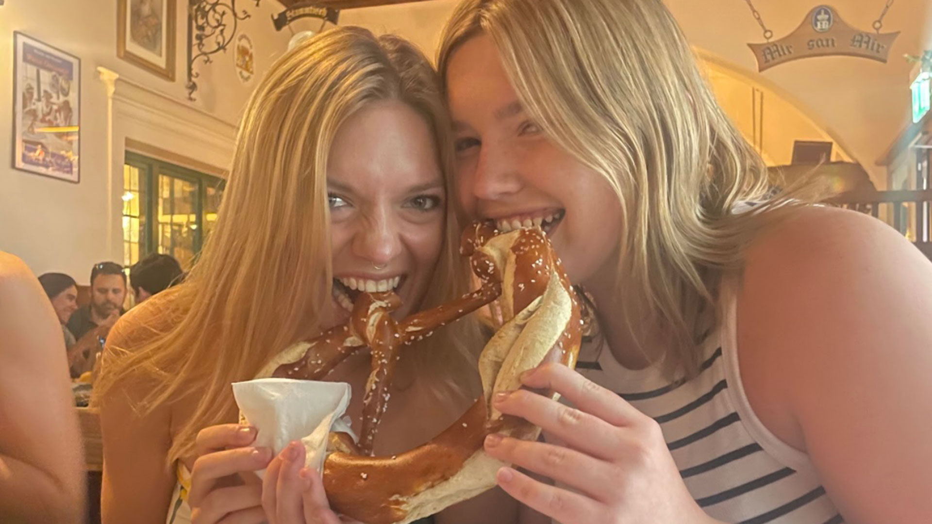 Two young women laughing while sharing and biting into a giant, salt-covered soft pretzel at an indoor restaurant.