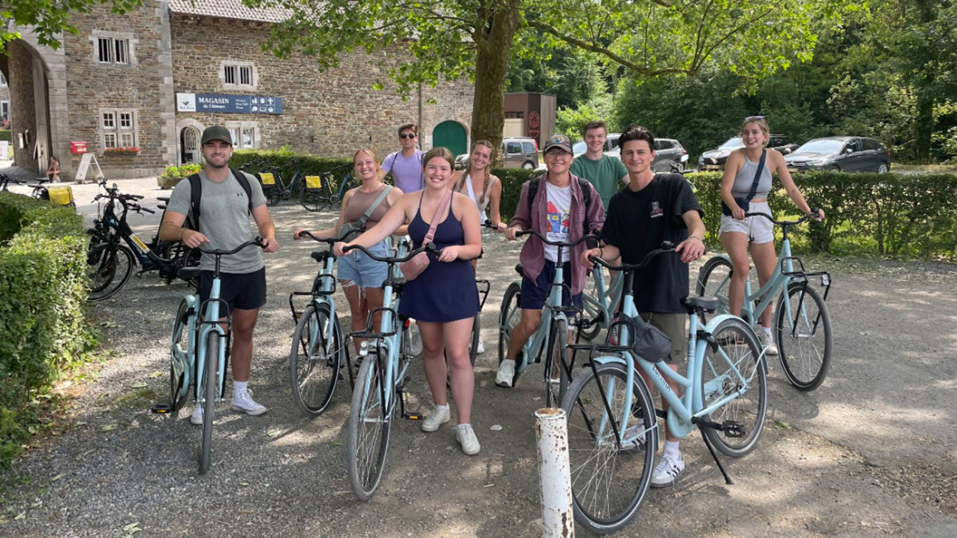 A group of smiling students posing with light blue bicycles in front of a rustic stone building on a sunny day.