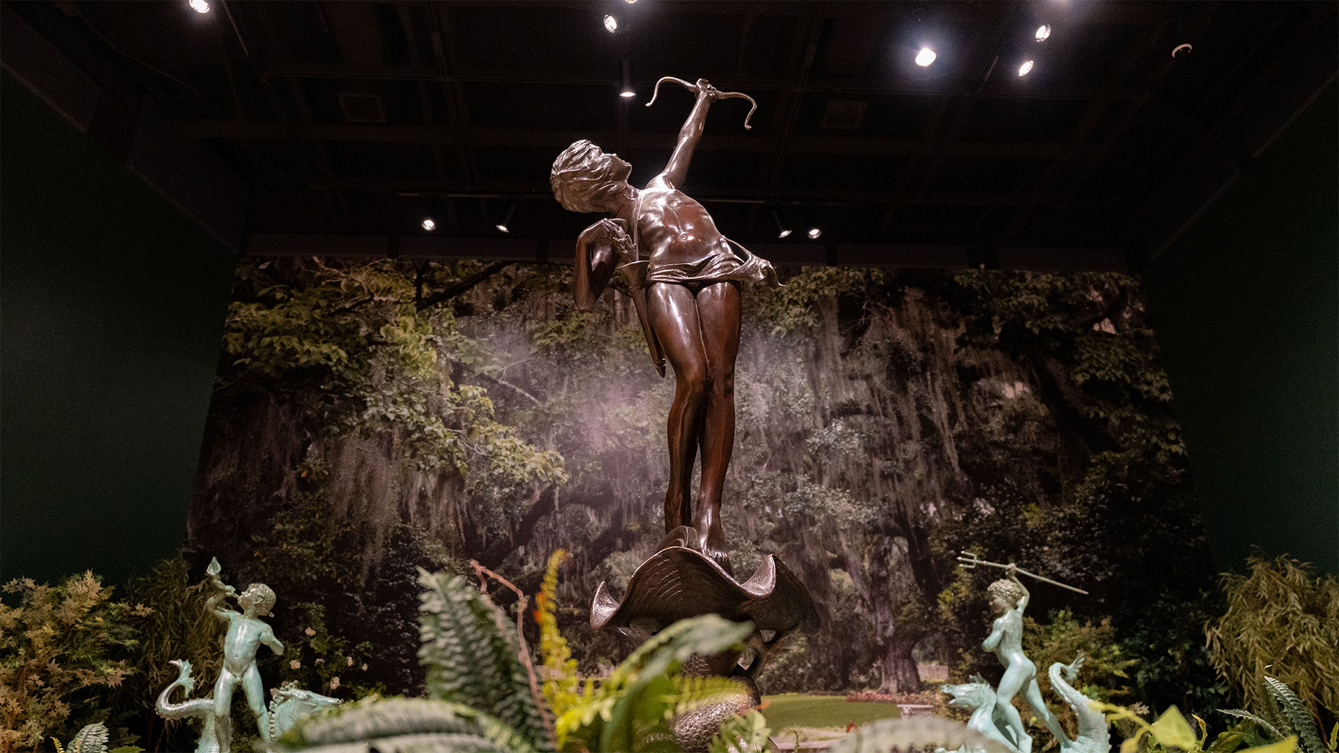 A bronze statue of a cherub points a bow and arrow skyward. The statue is surrounded by greenery.