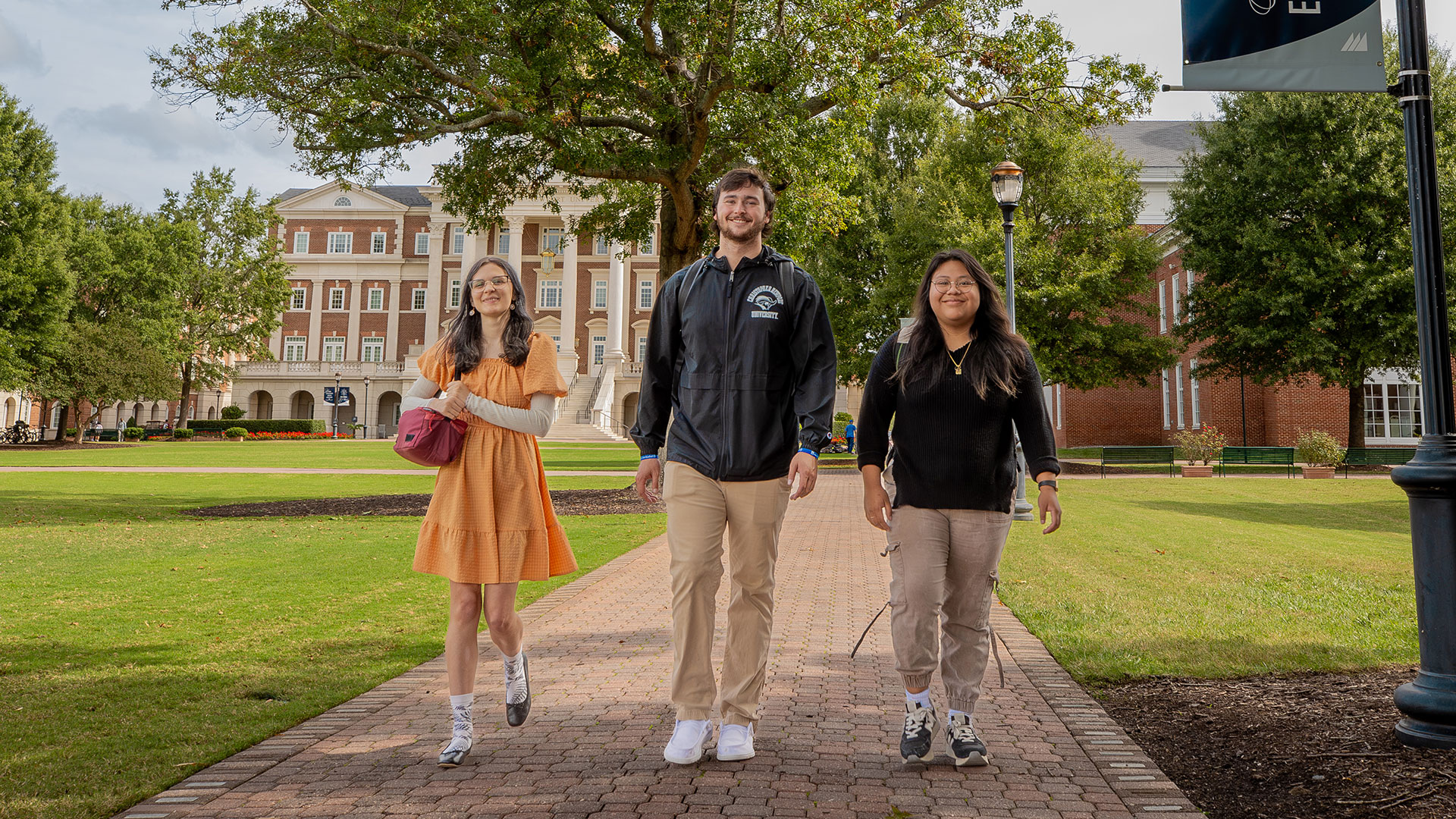  Three college students (two women and one man) walk together down a brick path on a university campus lawn toward a large brick administration building in the background.